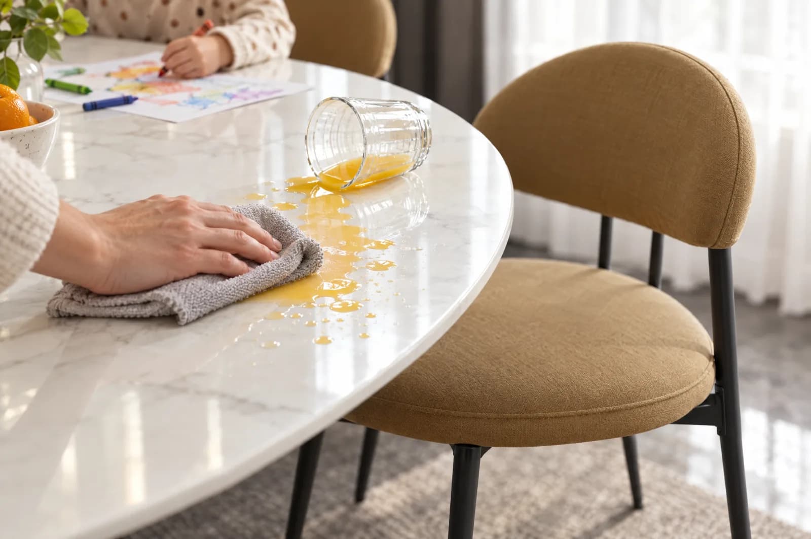 Hand wiping spilled orange juice off a white porcelain dining table with a cloth, beside a tan upholstered chair on black steel legs, with a child colouring at the table in the background