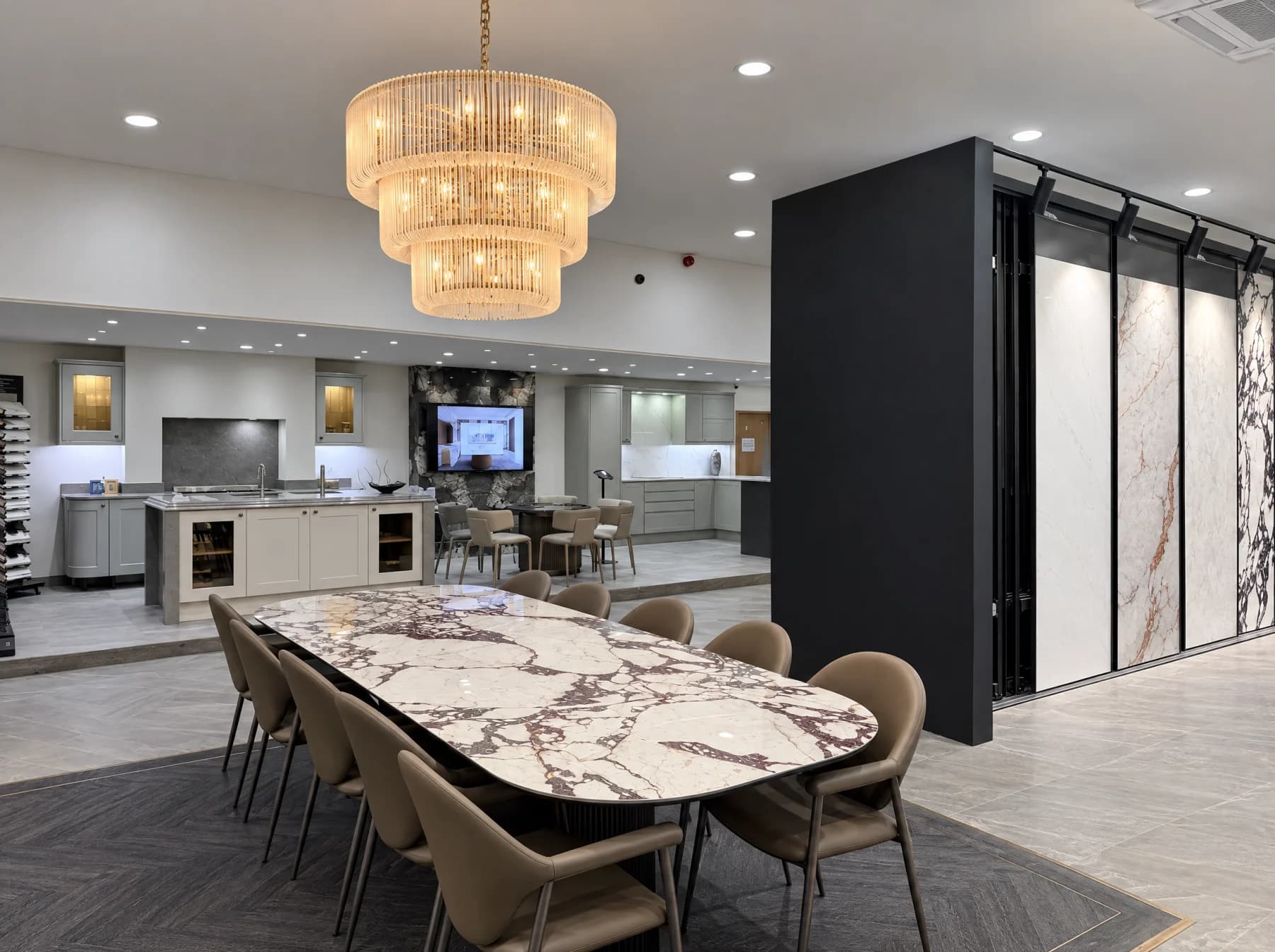 The BeMade showroom: a long oval porcelain dining table with burgundy veined marble-effect top and ten taupe chairs under a tiered chandelier, beside a backlit display wall of full size slabs